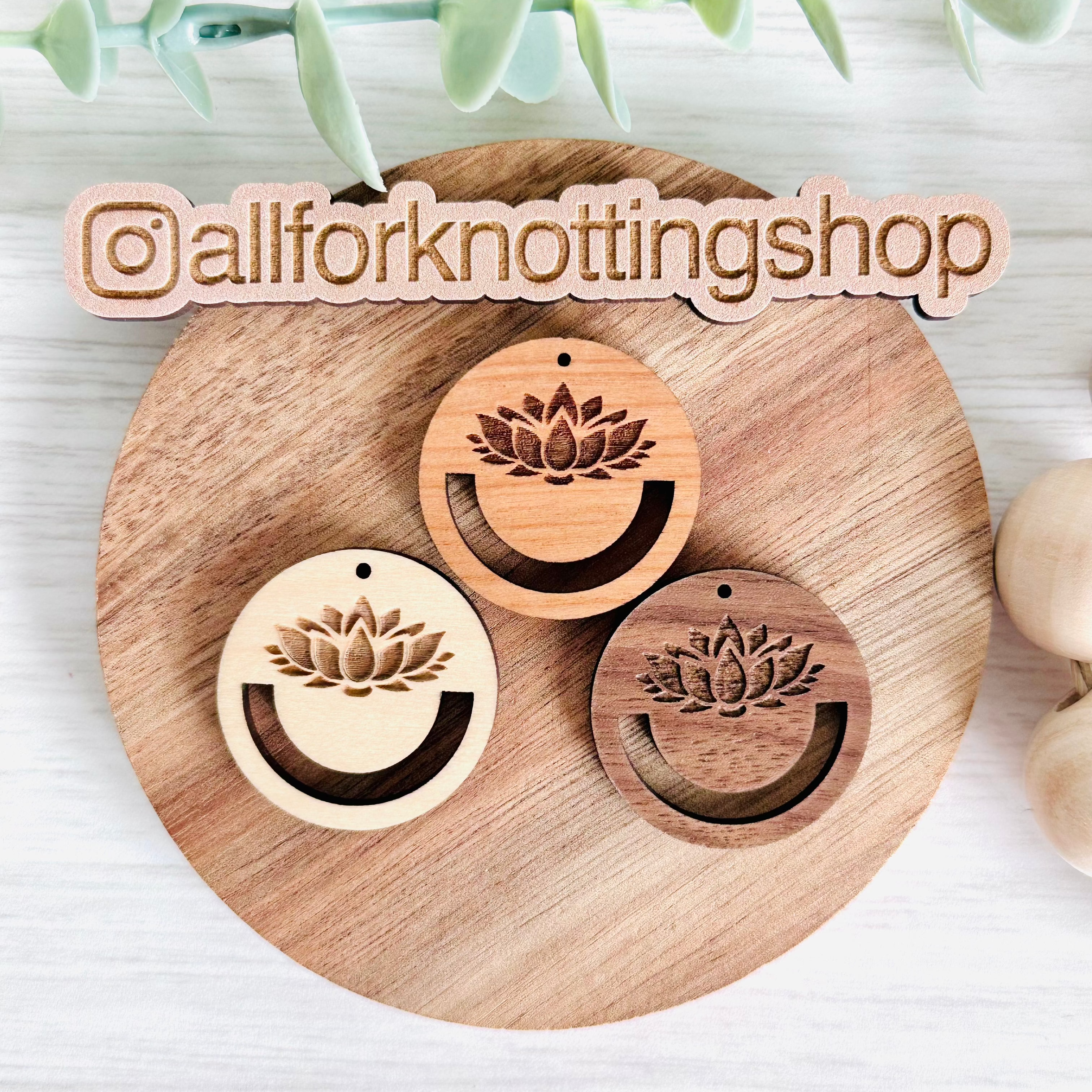 Three wooden earring blanks with a lotus design, presented on a wooden platter, with a logo and hashtag visible indicating the brand.